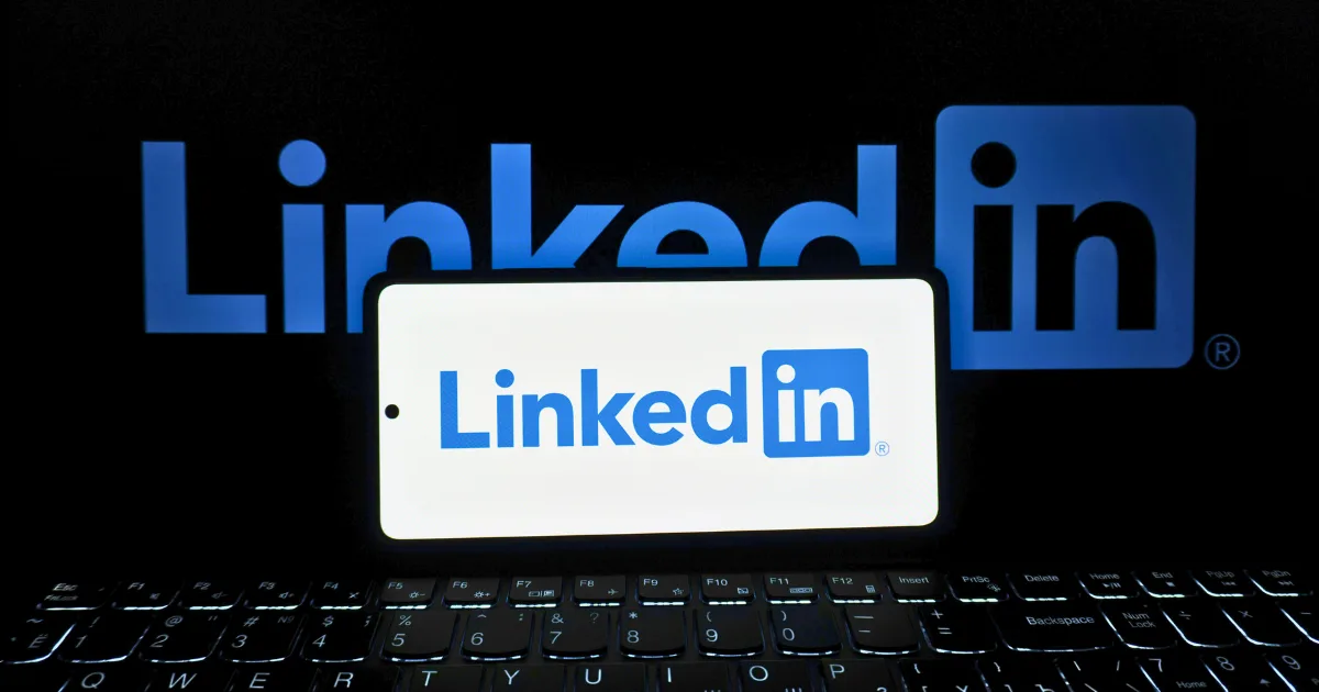 A smartphone displaying the LinkedIn logo sits on a keyboard, hinting at an effective LinkedIn content strategy, with a larger LinkedIn logo visible on a screen in the background.