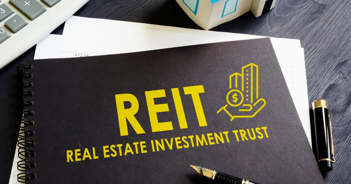A black notebook with REIT Real Estate Investment Trust and a building icon in yellow on the cover sits on a desk with a pen, calculator, and small house model—perfect for tracking your REIT investment strategies and financial planning.