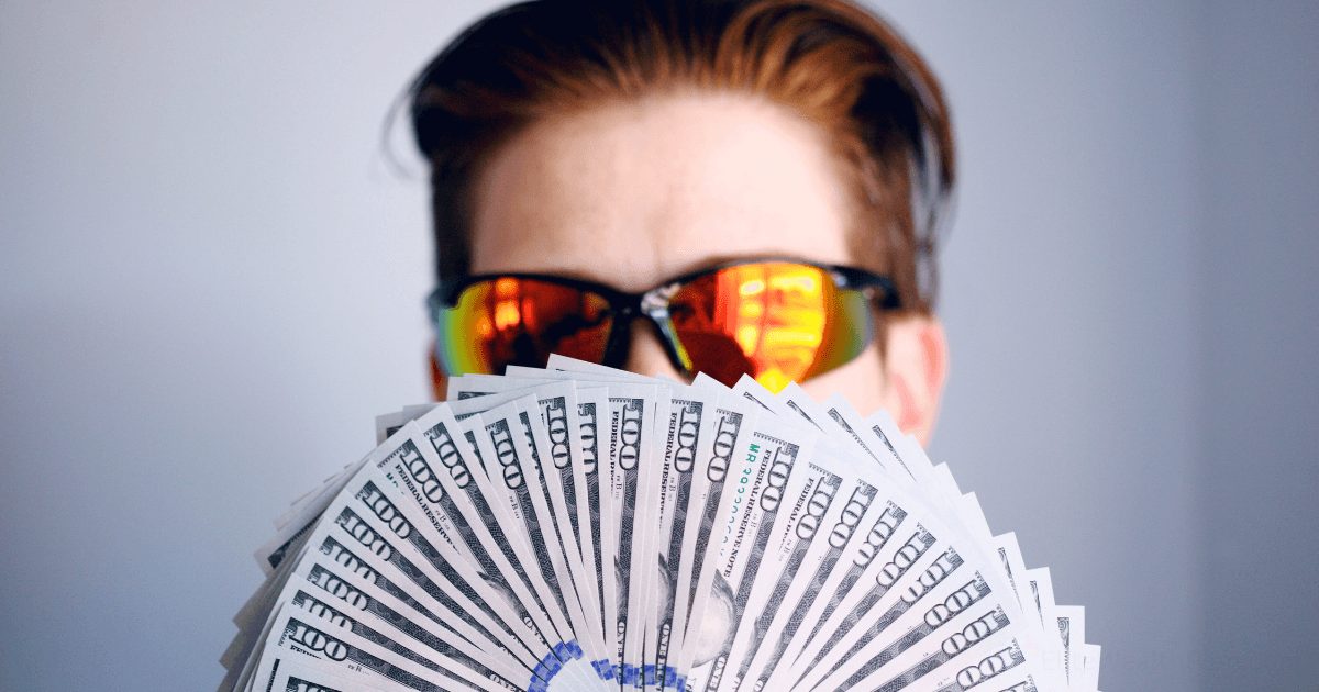A person wearing reflective sunglasses holds a large fan of U.S. hundred-dollar bills, partially covering their face, illustrating success in an affiliate marketing case study against a plain light background.