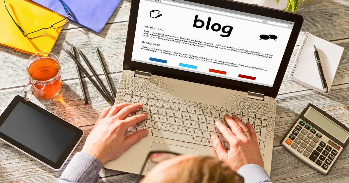 A person types on a laptop displaying a blog page, focusing on blog design elements. The desk also has a calculator, smartphone, notepad, pen, folder, glasses, and a cup of tea.
