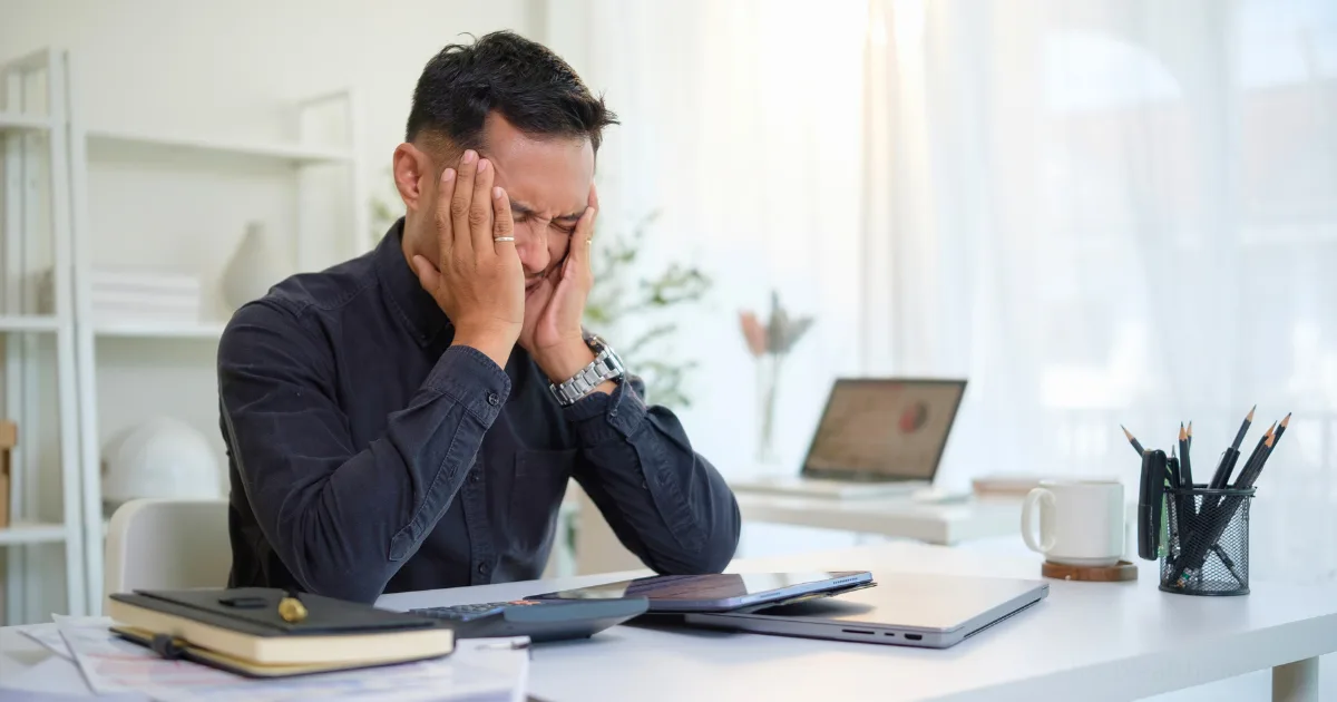A man sits at a desk with his eyes closed and hands covering his face, appearing stressed or frustrated—perhaps in need of a burnout recovery plan. The workspace includes a laptop, documents, and pens, with bright light streaming from the window behind him.