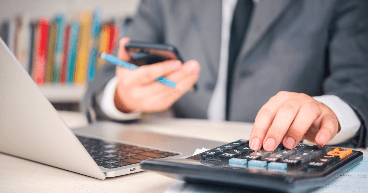 A person in a suit uses a calculator and smartphone while working at a desk with an open laptop, efficiently managing finances with a business expense tracking system; bookshelves are blurred in the background.