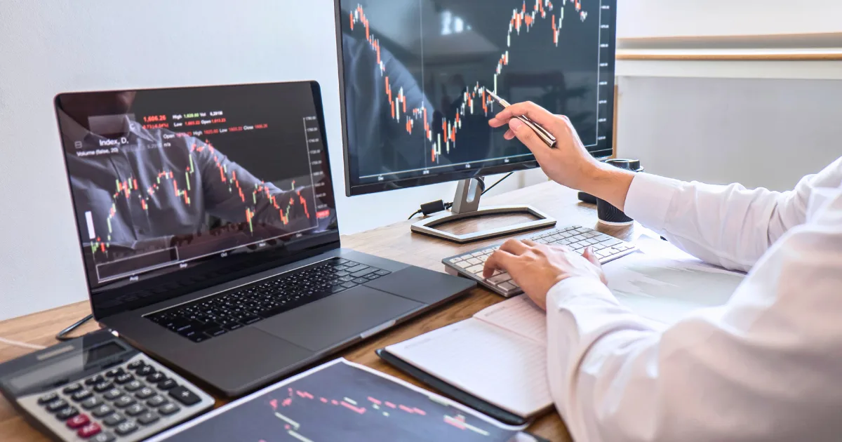 A person analyzes financial charts on a laptop and monitor at a desk with a keyboard, notepad, calculator, and documents, illustrating day trading for side income or in-depth stock market data analysis.