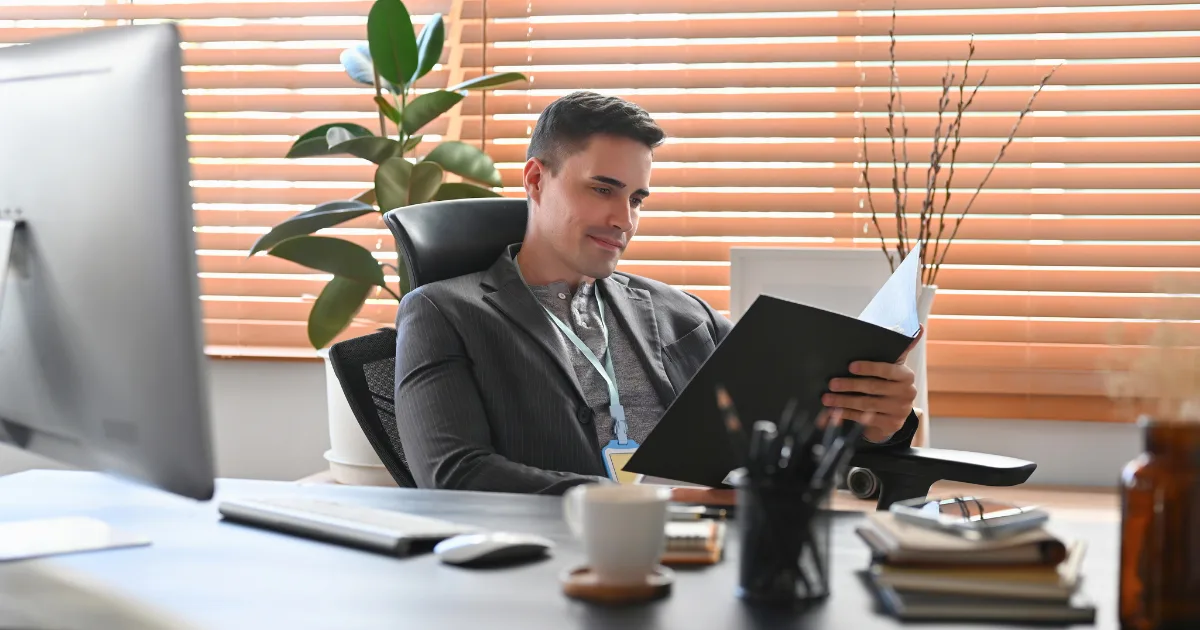 A man in a suit sits at a desk in an office, reading a document. A computer monitor, coffee cup, and office supplies are on the desk, suggesting he’s reviewing a decision-making framework. Blinds and a plant are visible in the background.