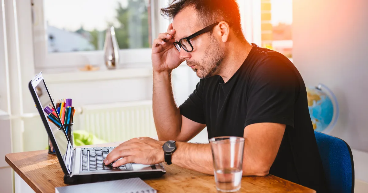 A man wearing glasses and a black t-shirt sits at a desk, looking intently at a laptop as he follows a deep work protocol. He appears focused or concerned, with one hand touching his forehead. A glass of water and office supplies are on the desk.