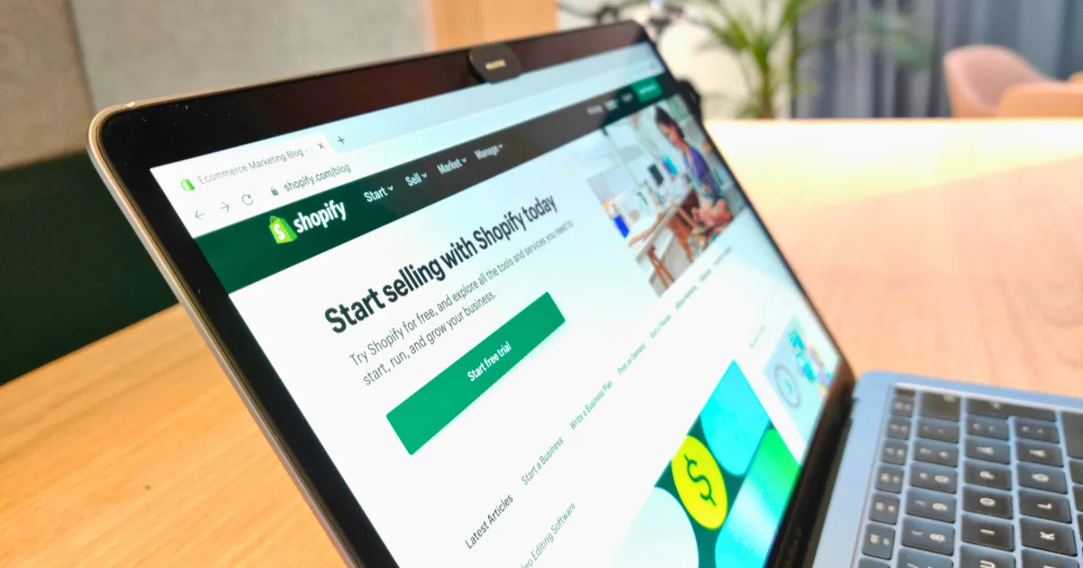 A laptop on a wooden desk displays the Shopify website homepage, inviting users to start selling with Shopify today. The scene hints at how digital product marketplaces can easily be managed from a modern workspace, with a blurred plant and office furniture in the background.