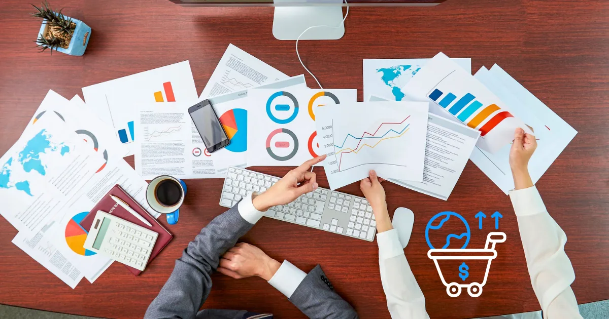 Two people at a desk review colorful charts and graphs, using a computer to analyze e-commerce analytics; coffee and a plant are nearby, with a shopping cart and growth icons in the foreground.