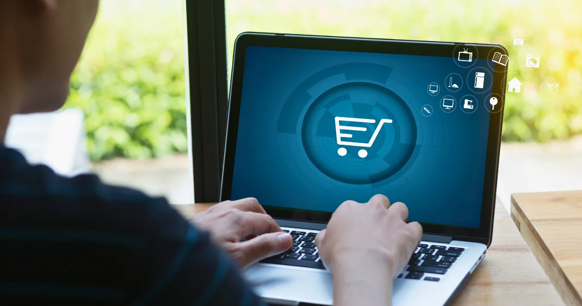 A person using a laptop with an online shopping cart icon displayed on the screen, highlighting e-commerce marketing plan strategies, as sunlight streams in through a window in the background.