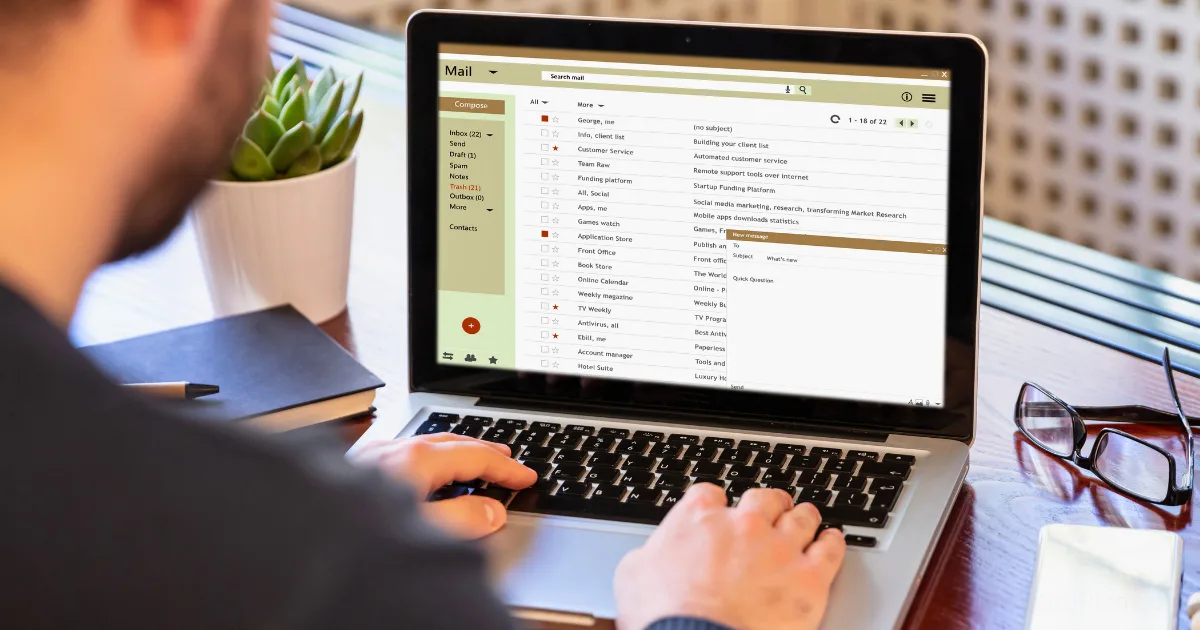 A person types on a laptop displaying an email inbox, focused on email list building. A notebook, pen, glasses, and a small potted plant are neatly arranged on the wooden desk nearby.