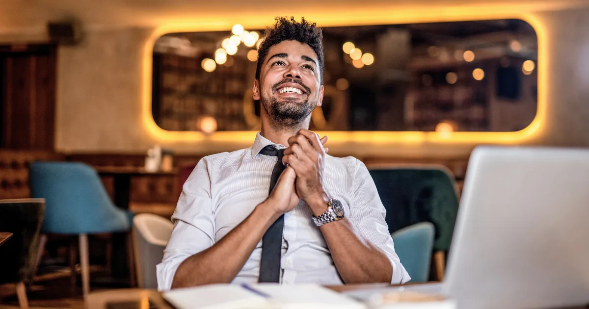 A man in a white shirt and tie sits at a table, smiling and looking upward with hands clasped, embodying the journey from failure to success. An open laptop is in front of him, with warm ambient lighting softly illuminating the background.