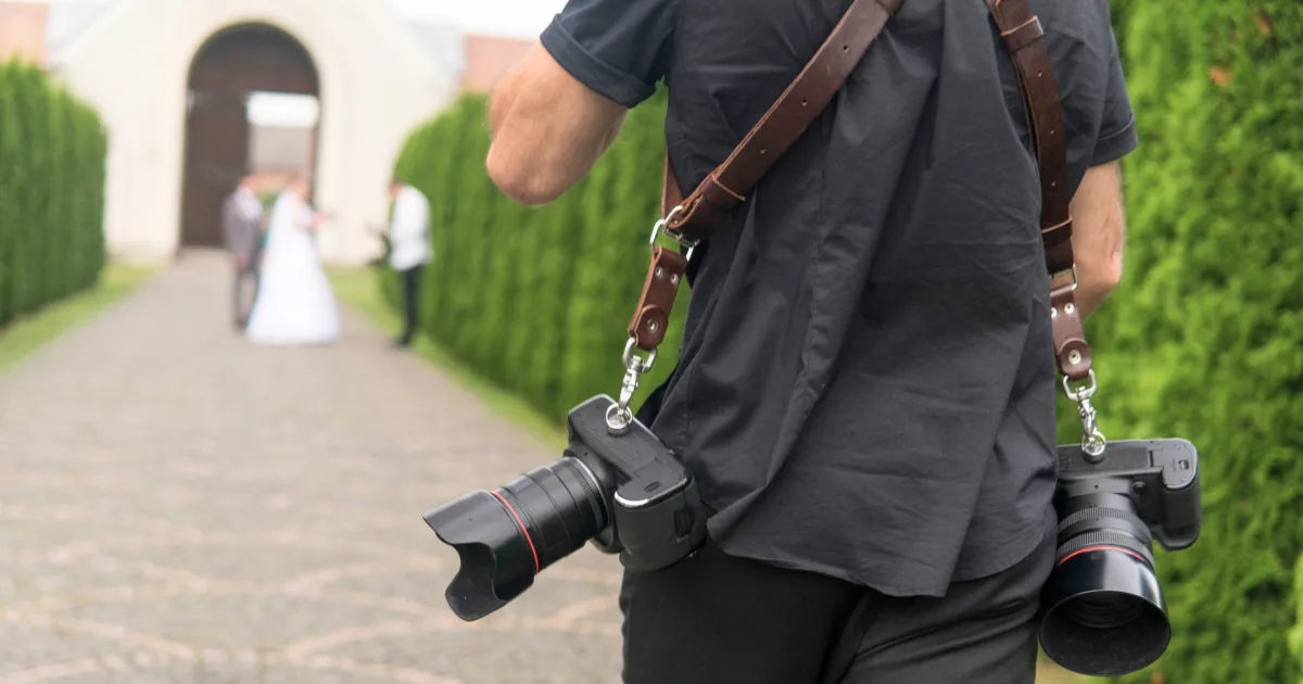 A photographer specializing in event photography, wearing a black shirt and camera harness with two cameras, walks on a cobblestone path toward a bride and groom standing together in the distance, with greenery lining the walkway.