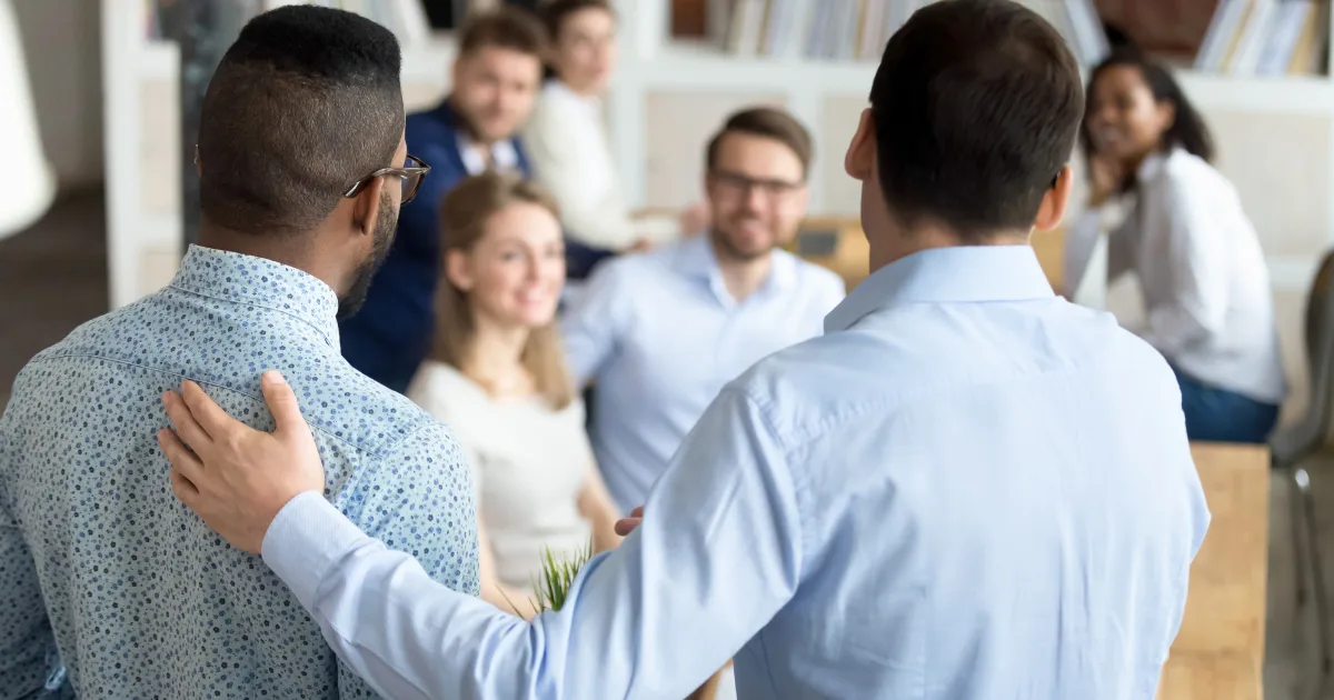 Two men stand with their backs to the camera—one with his arm around the other’s shoulders, perhaps celebrating a first hire—as they face a group of smiling colleagues in a well-lit, casual office setting.