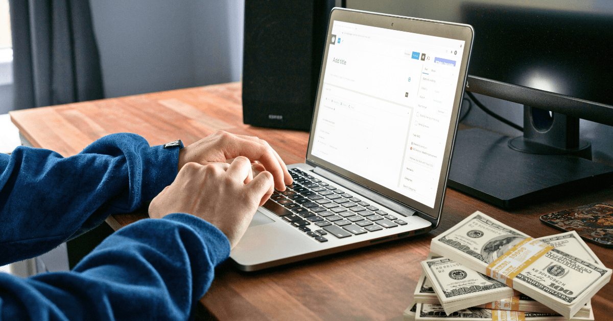 A person wearing a blue sweatshirt types on a laptop at a wooden desk, surrounded by stacks of hundred-dollar bills—an inspiring scene for anyone dreaming of making money in their first year blogging. A monitor and speaker complete the setup.