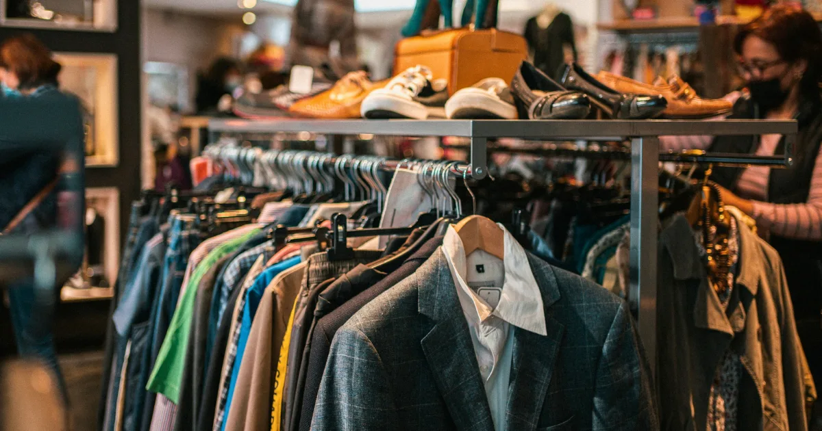 A clothing rack filled with assorted shirts, jackets, and blazers in a store. Shoes and bags are displayed on the shelf above as shoppers browse, flipping through stylish finds in the warm, busy scene.
