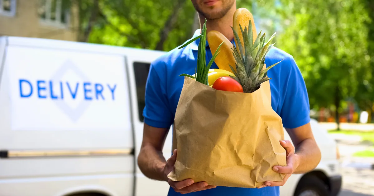 A person in a blue shirt holds a paper bag filled with groceries, including a pineapple and baguettes, standing in front of a food delivery van on a sunny day.