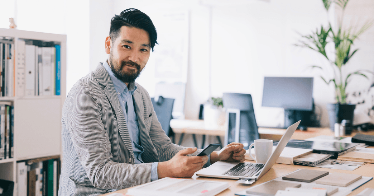 A man in a light suit jacket sits at a desk with a laptop, holding a smartphone. He looks at the camera and smiles, confident in building future-proof income streams. The modern office has shelves, plants, and computer monitors in the background.