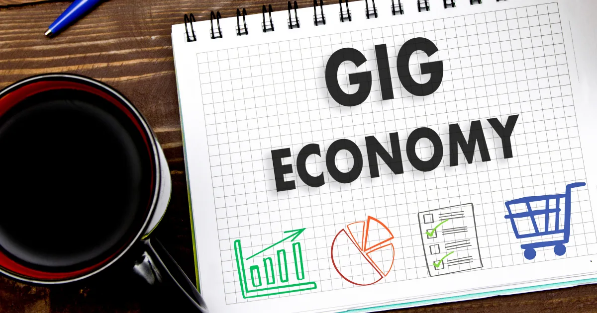 A notebook with “GIG ECONOMY” written on a grid page, surrounded by colorful icons of a bar graph, pie chart, checklist, and shopping cart—perfect for planning your next move with gig economy apps. A cup of coffee and blue pen rest on the wooden surface.
