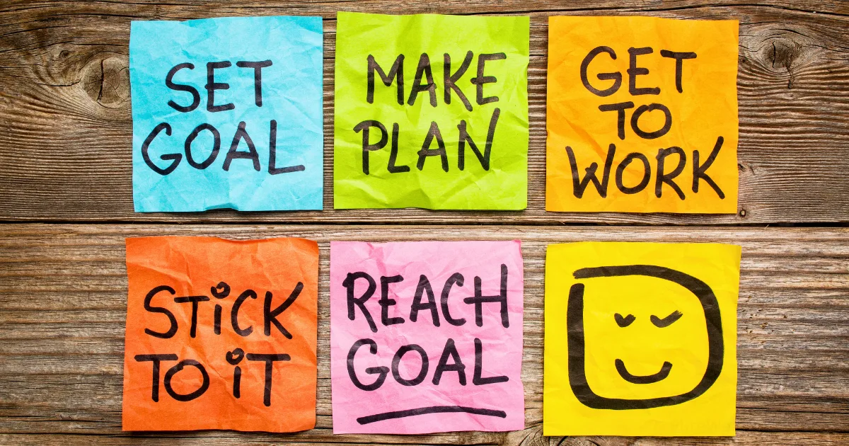 Six colorful sticky notes on a wooden surface illustrate a goal setting framework, each with motivational messages: SET GOAL, MAKE PLAN, GET TO WORK, STICK TO IT, REACH GOAL, and a smiling face drawing.