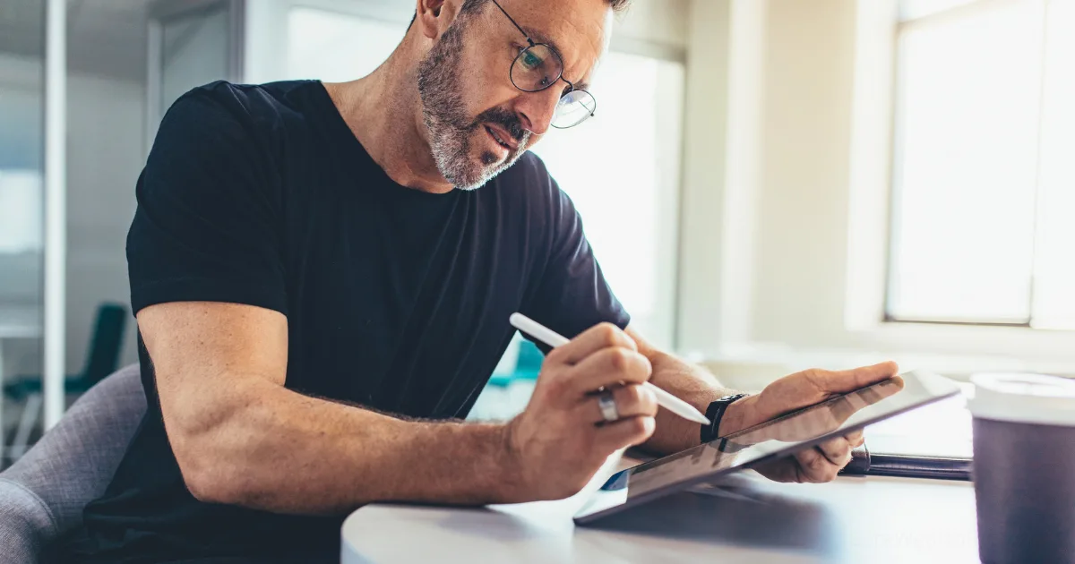 A man wearing glasses and a black t-shirt sits at a desk, using a stylus to write or draw on a tablet—perhaps planning habit stacking for entrepreneurs. Focused, he works beside a coffee cup as sunlight streams through the windows behind him.