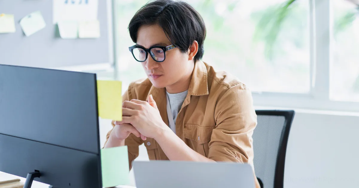 A person wearing glasses and a brown shirt sits at a desk, looking intently at a computer monitor with sticky notes attached—perhaps exploring an imposter syndrome toolkit—with a laptop nearby and bright windows in the background.