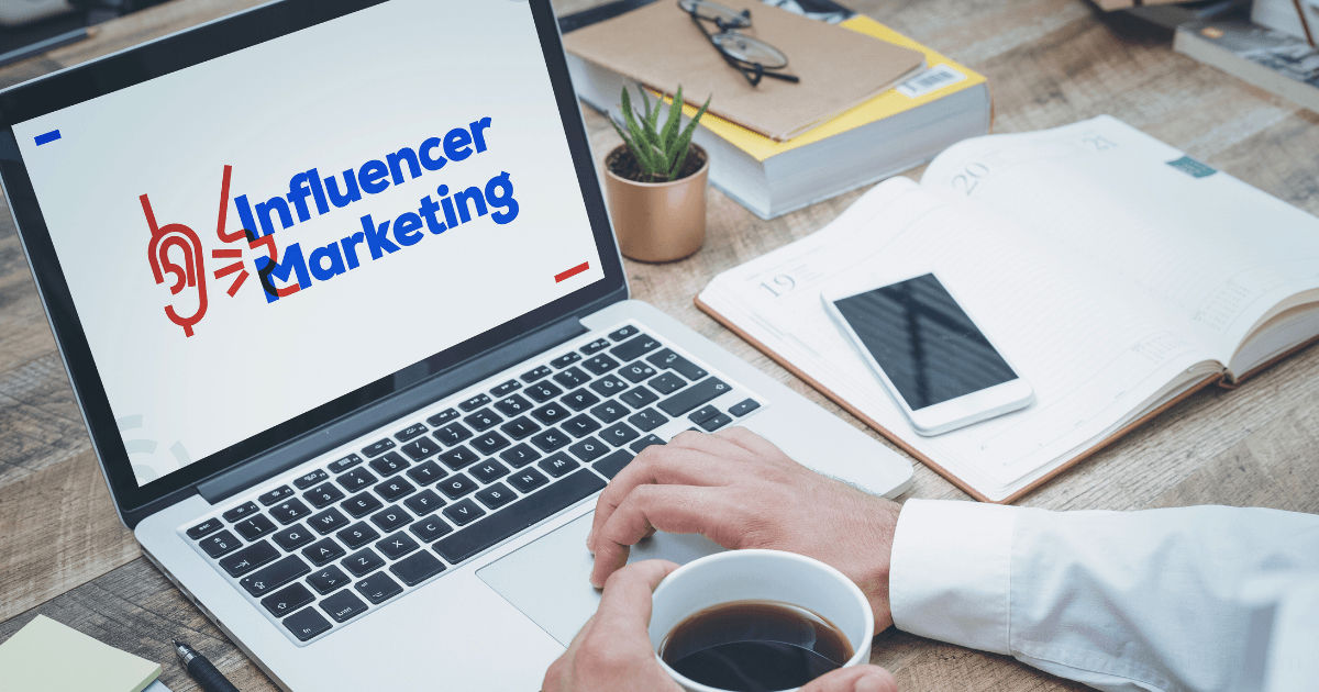 A person holding a coffee cup works at a desk with a laptop displaying influencer marketing strategies. Nearby are a smartphone, notebook, pen, and folders.