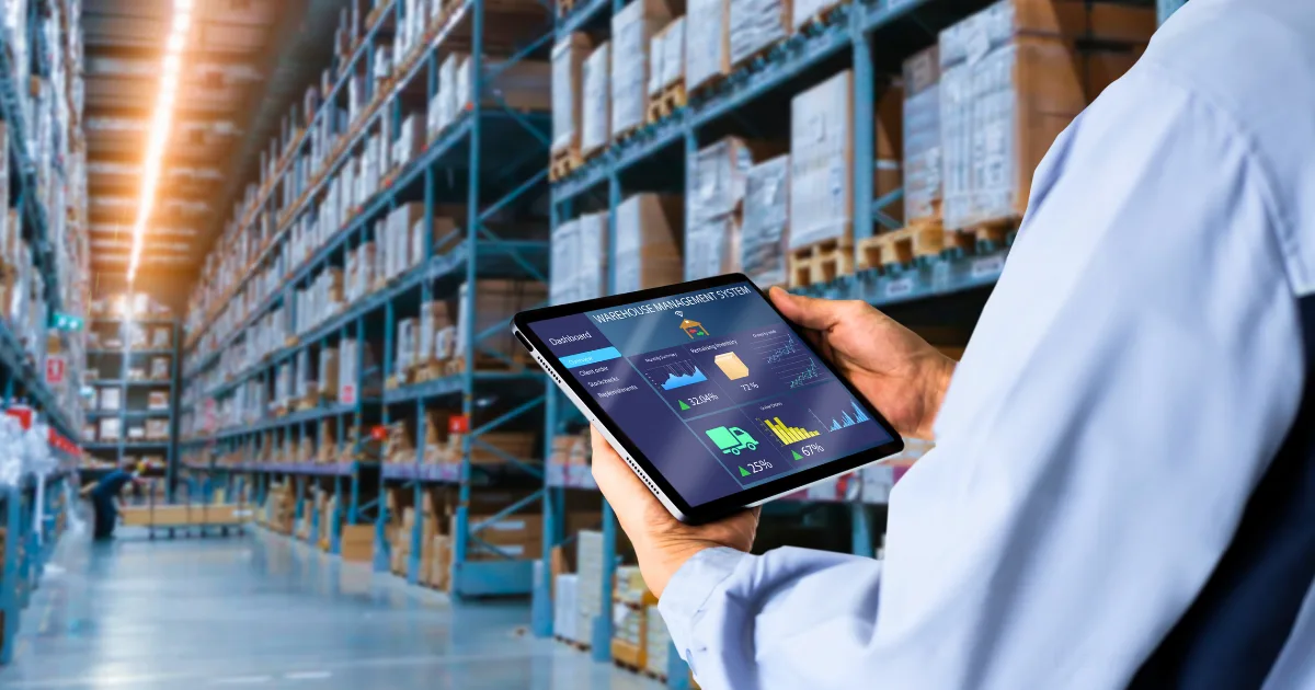 A person in a warehouse holds a tablet displaying inventory management for solopreneurs, with tall shelves of boxes and products stacked in the background.