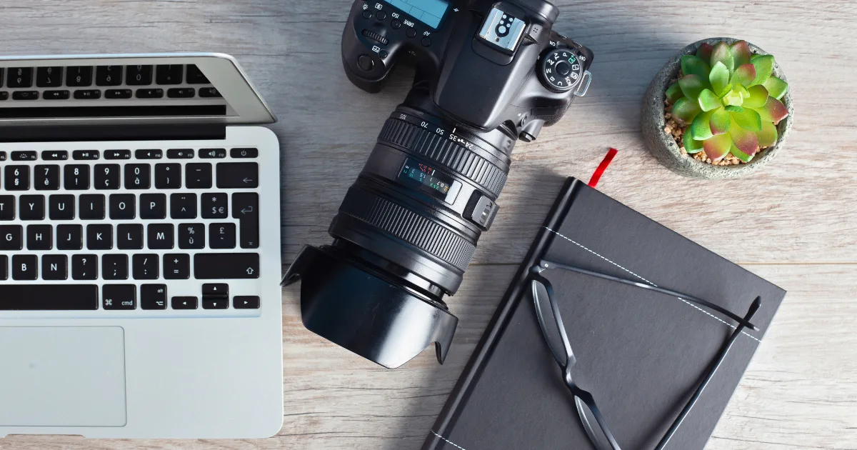 A camera, a laptop, a black notebook with glasses and a red pen, and a small potted succulent are arranged on a wooden desk—perfect for planning projects or licensing your work.