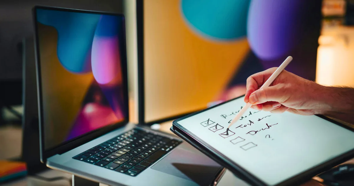 A person uses a micro-task strategy, writing a checklist on a tablet with a stylus, while a laptop and two monitors displaying colorful abstract backgrounds complete the workspace.
