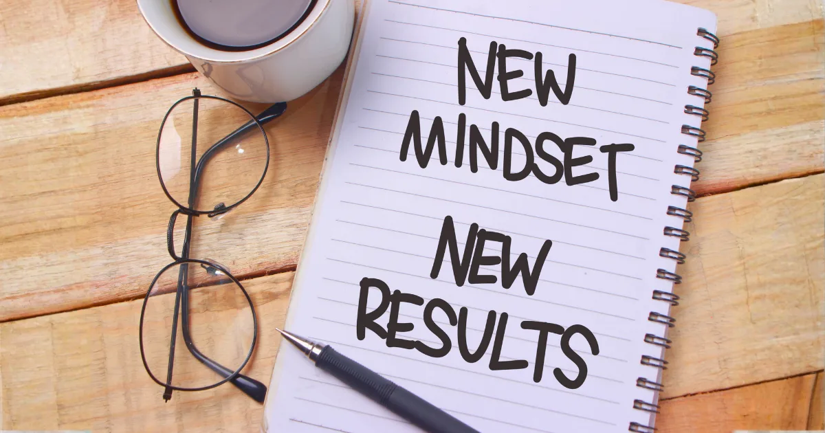 A spiral notebook with the words NEW MINDSET NEW RESULTS written on lined pages, illustrating powerful mindset shifts, sits next to a pair of glasses, a pen, and a cup of coffee on a wooden surface.