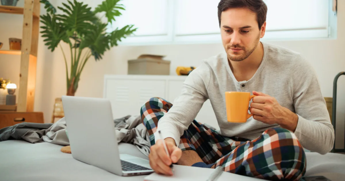 A man in pajamas sits on a bed with a laptop, holding an orange mug in one hand and writing with the other—capturing his morning routine experiment. A plant and shelves fill the background as natural light streams through the window.
