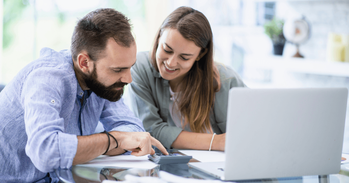 A man and a woman sit at a table with a laptop, smiling and looking at paperwork while the man uses a calculator—reviewing their personal finance system for entrepreneurs in a bright home office.