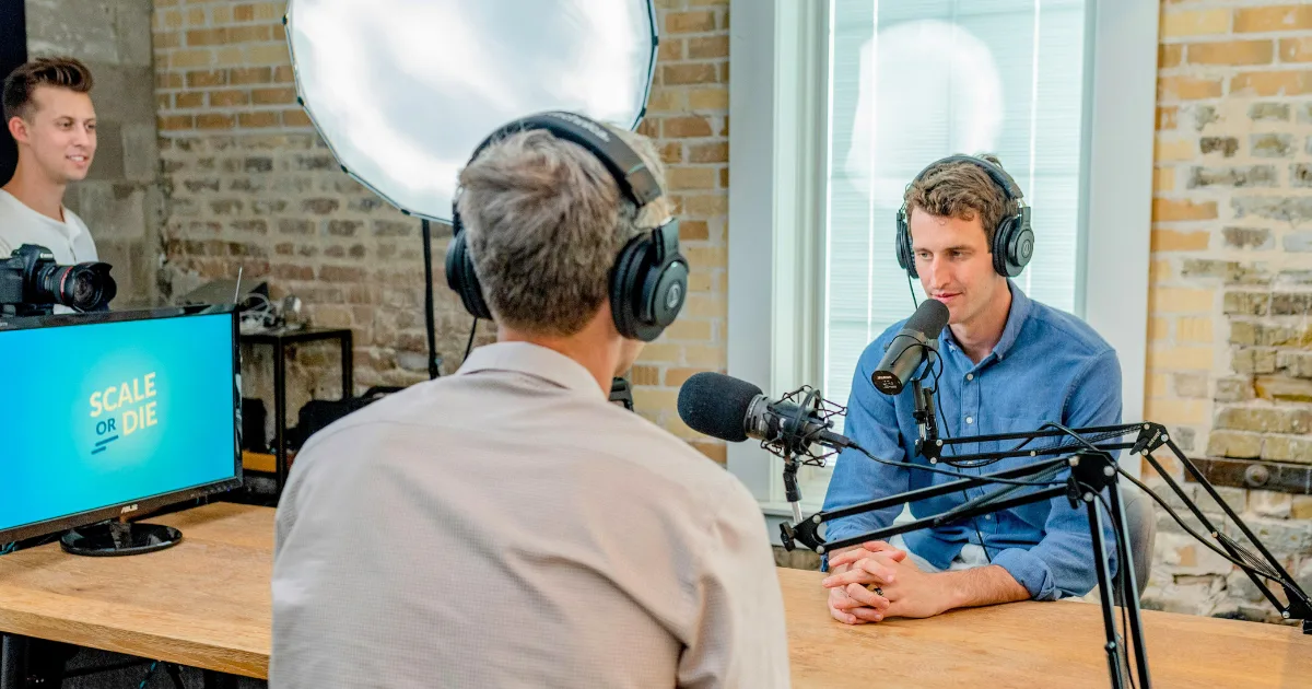 Two men wearing headphones speak into microphones during a podcast launch in a studio, while a third person stands nearby. A bright light and a monitor displaying SCALE OR DIE are visible in the background.