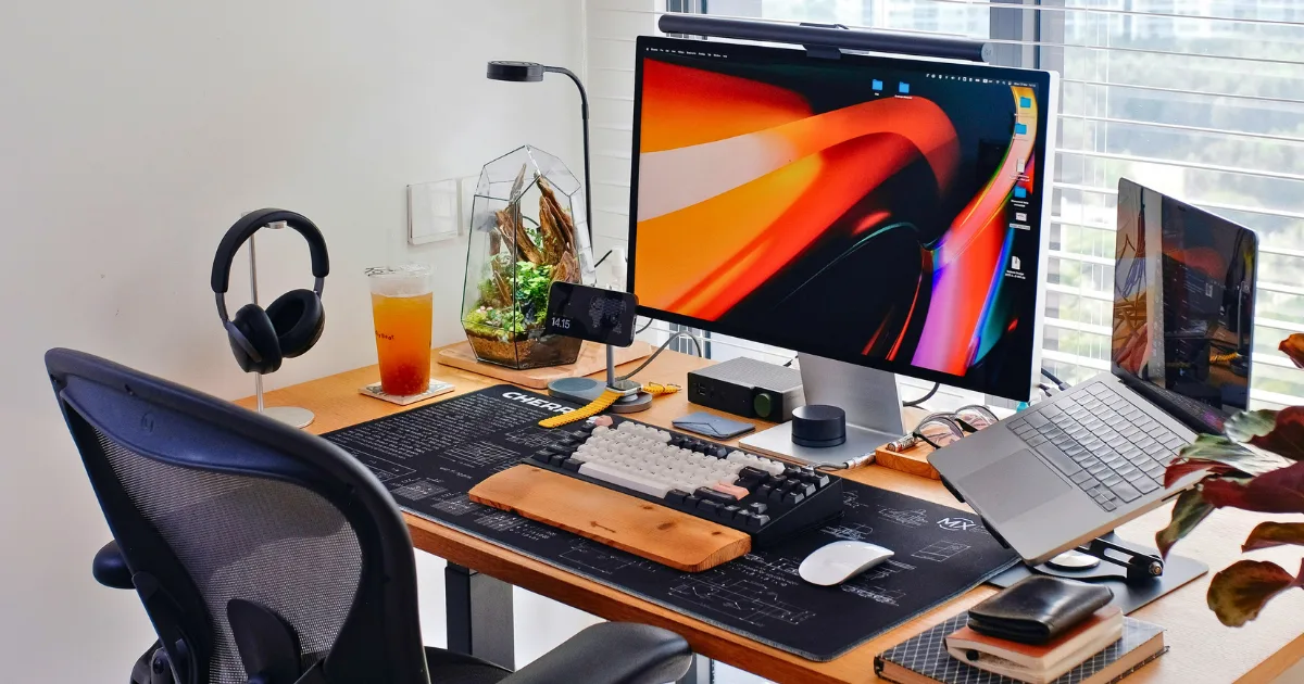 A tidy workspace featuring productivity hardware like a desktop monitor, laptop on a stand, wireless keyboard, headphones, mouse, drink, lamp, potted plant, and office supplies arranged on a wooden desk by a window with blinds.