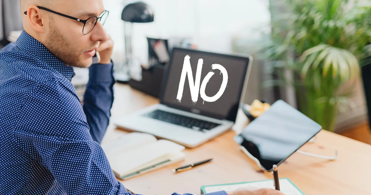 A man in glasses sits at a desk with a laptop displaying the word NO in bold white letters—a scene illustrating rejection resilience. The workspace includes scattered papers, a tablet, and a large green plant in the background.