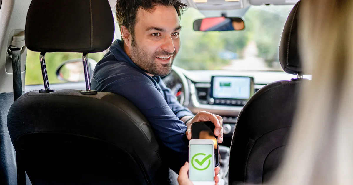 A smiling man in a rideshare driving seat looks back at a passenger, who is holding up a smartphone displaying a large green checkmark on the screen.