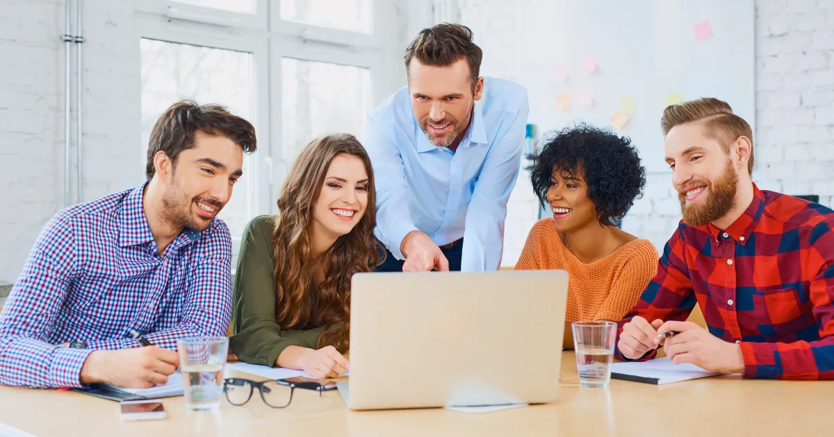 Five people sit at a table in a bright, modern office with large windows, smiling and looking at a laptop screen together—brainstorming ideas to scale your agency. Three have notebooks open, and glasses of water are on the table.