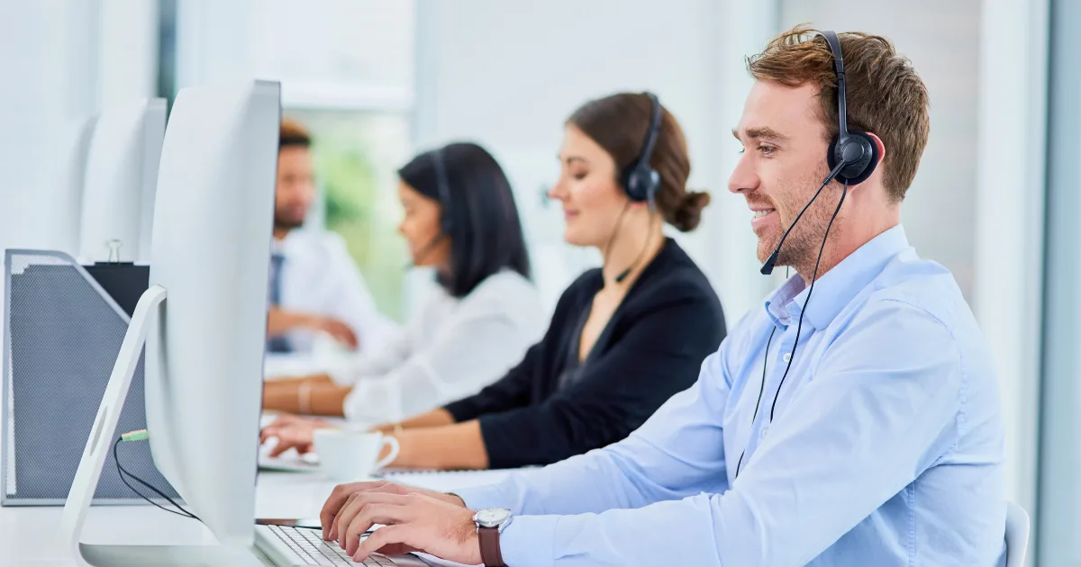 Four people sit at desks wearing headsets and working on computers in a bright, modern office. The focus is on a smiling man in the foreground, illustrating a team dedicated to scaling customer support, with colleagues blurred in the background.