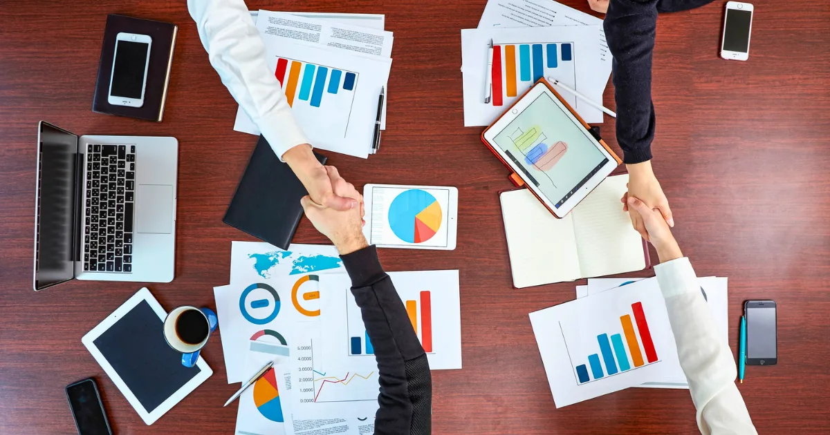 Overhead view of a wooden table with laptops, tablets, and documents displaying charts and template business forms. Three people are shaking hands across the table, suggesting a business meeting or agreement.