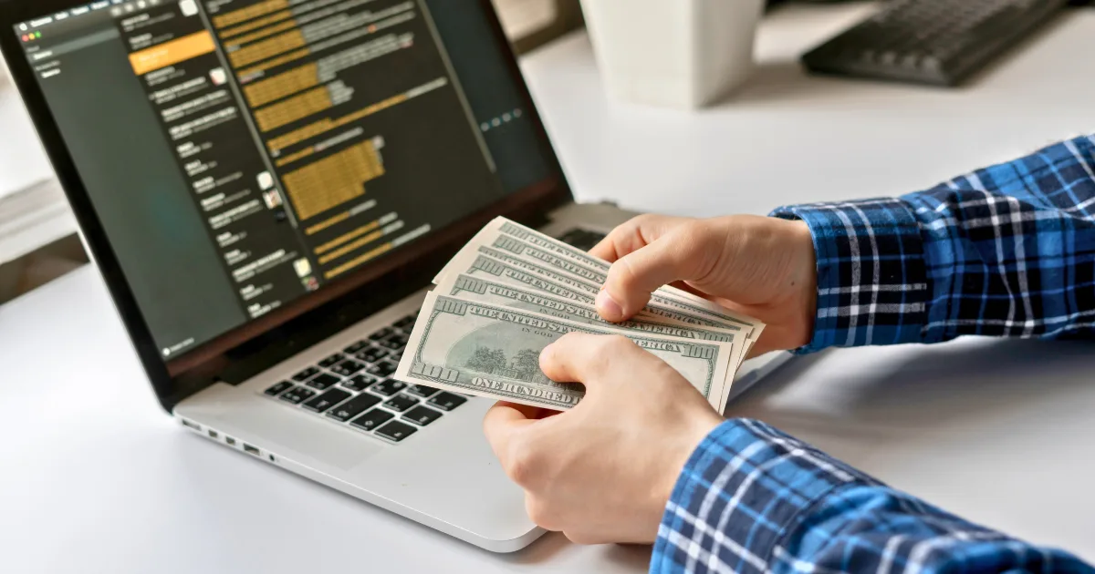 A person wearing a blue plaid shirt counts several US $100 bills while sitting at a desk with an open laptop displaying emails, reflecting on their journey from side hustle to full-time success.