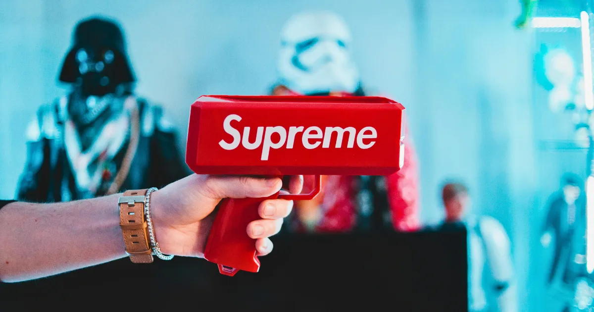 A person holds a red Supreme cash gun in the foreground—perfect for flexing your sponsored content rate—as blurry figures of Darth Vader and a Stormtrooper appear in the blue-lit background.