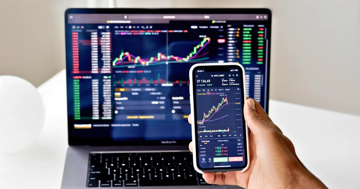 A person holds a smartphone displaying a stock chart in front of a laptop with similar financial data, suggesting they are monitoring trends and possibly refining their stock market entry plan.