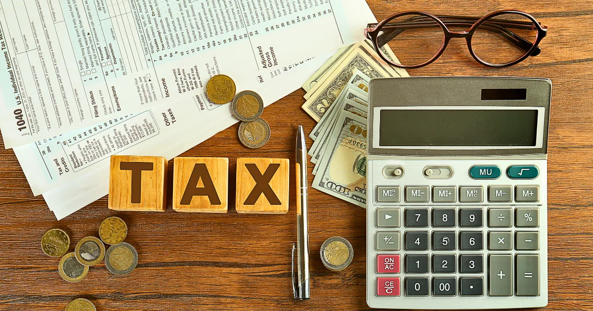 A calculator, pen, eyeglasses, U.S. dollar bills and coins, a tax planning calendar, and wooden blocks spelling TAX sit on top of tax forms on a wooden desk.