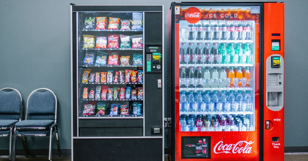 A snack vending machine and a Coca-Cola drink vending machine, part of a thriving vending machine business, stand side by side against a gray wall, next to a row of gray chairs.