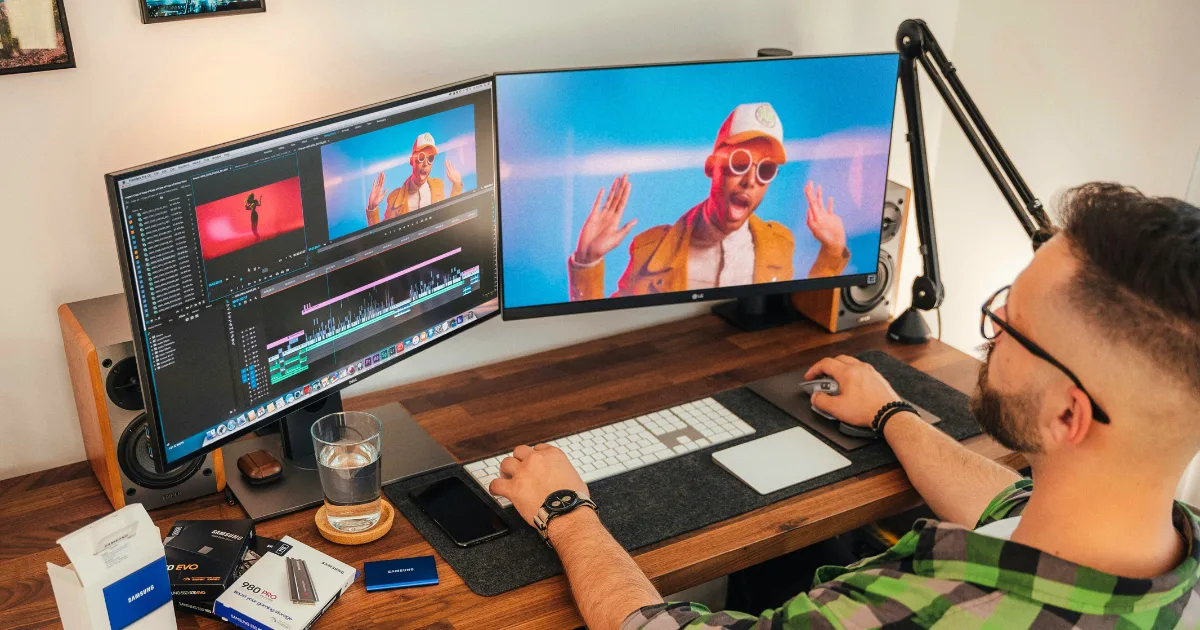 A person sits at a desk using two large monitors and video editing shortcuts to edit a colorful video of a performer in sunglasses and a hat; the workspace has a keyboard, mouse, water glass, and tech accessories.