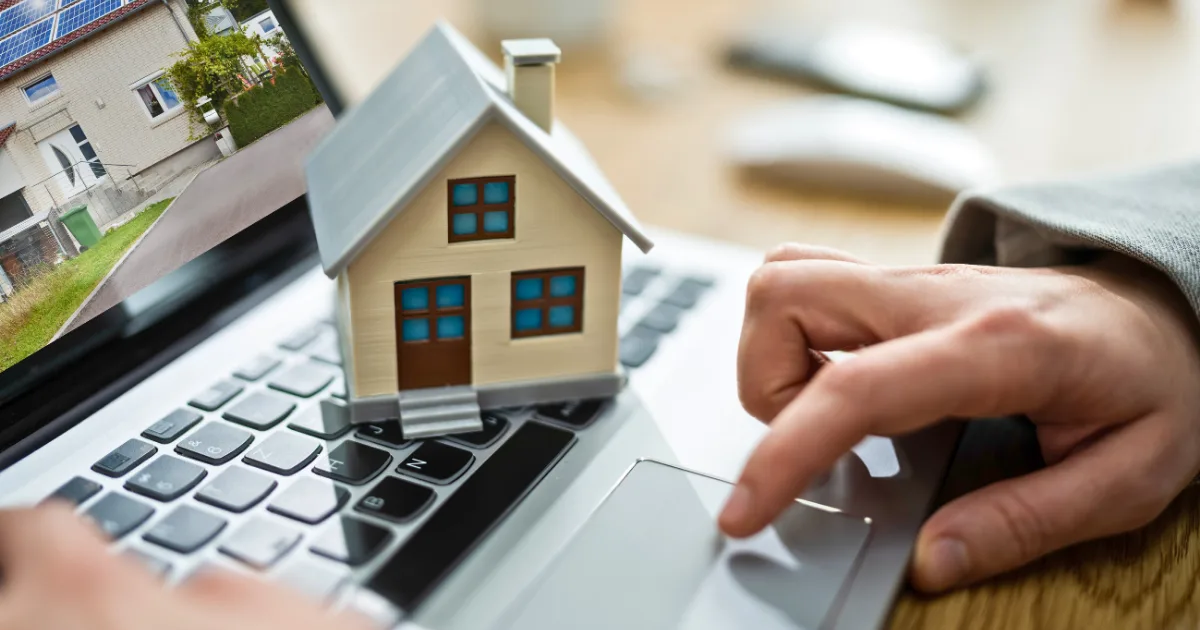 A person uses a laptop with a small model house placed on the keyboard, suggesting virtual real estate browsing or digital property management.