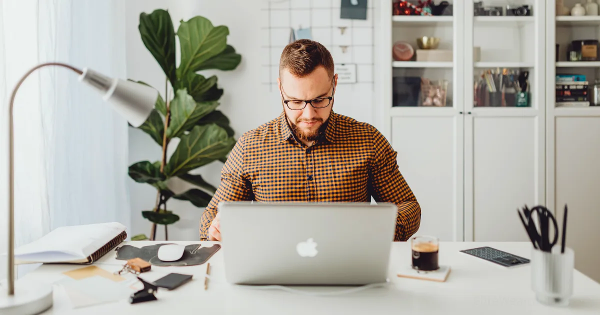 A man wearing glasses and a checked shirt sits at a white desk, working on his laptop with work-from-home discipline. The desk holds a notebook, lamp, coffee, and plants, while shelves and a large plant appear in the background.