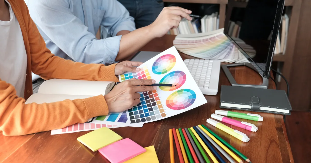 Two people at a desk reviewing color swatches and color wheels, surrounded by colored pencils, sticky notes, and a keyboard—an inviting scene perfect for exploring graphic design for non-designers in a creative workspace.