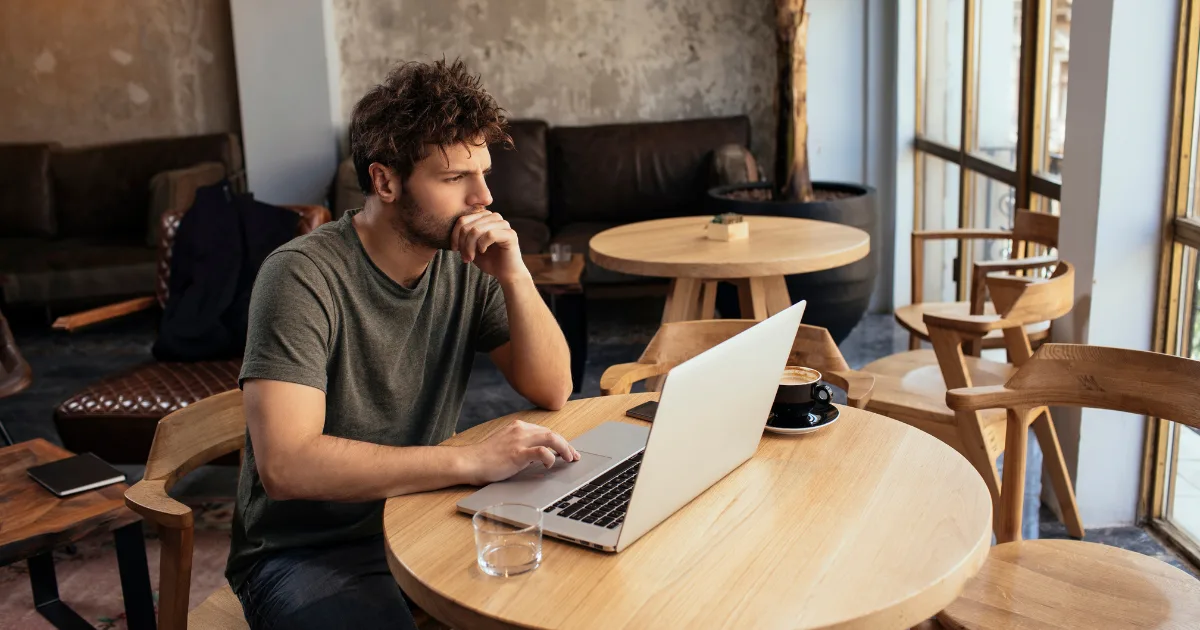 A man sits at a round wooden table in a cafe, looking thoughtfully at his open laptop as he researches pricing psychology. He rests his chin on his hand, with a cup of coffee and a glass of water beside him. The cafe glows with natural light.