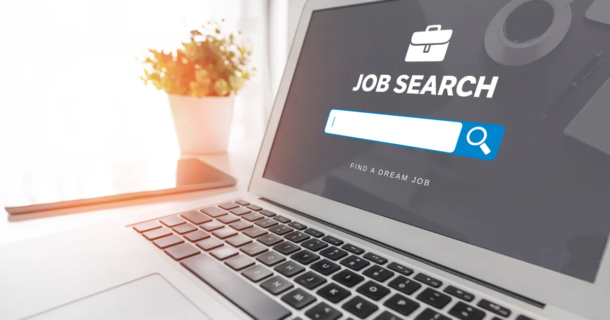 A laptop displays a job search webpage featuring remote job boards with a search bar and briefcase icon. A potted plant sits in the background on a white desk, illuminated by natural light.