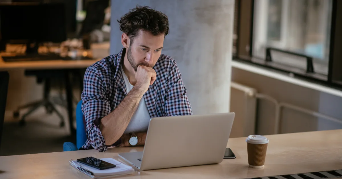 A man with a beard sits at a desk, looking thoughtfully at an open laptop—perhaps planning his social media management business. A smartphone, notebook, and takeaway coffee cup rest on the softly lit office desk.